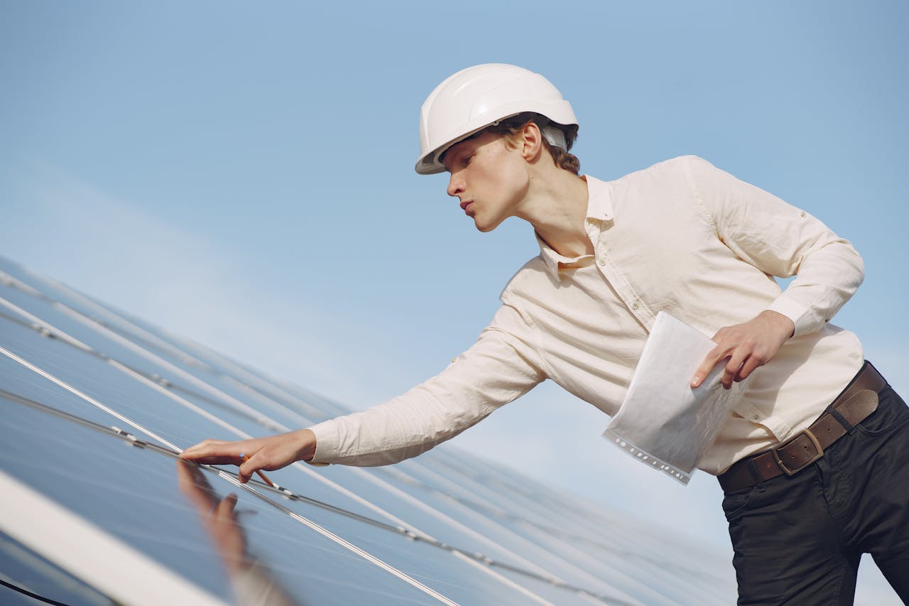 Low angle of young male inspector with documents in formal wear and hardhat standing against solar panels and checking details of construction